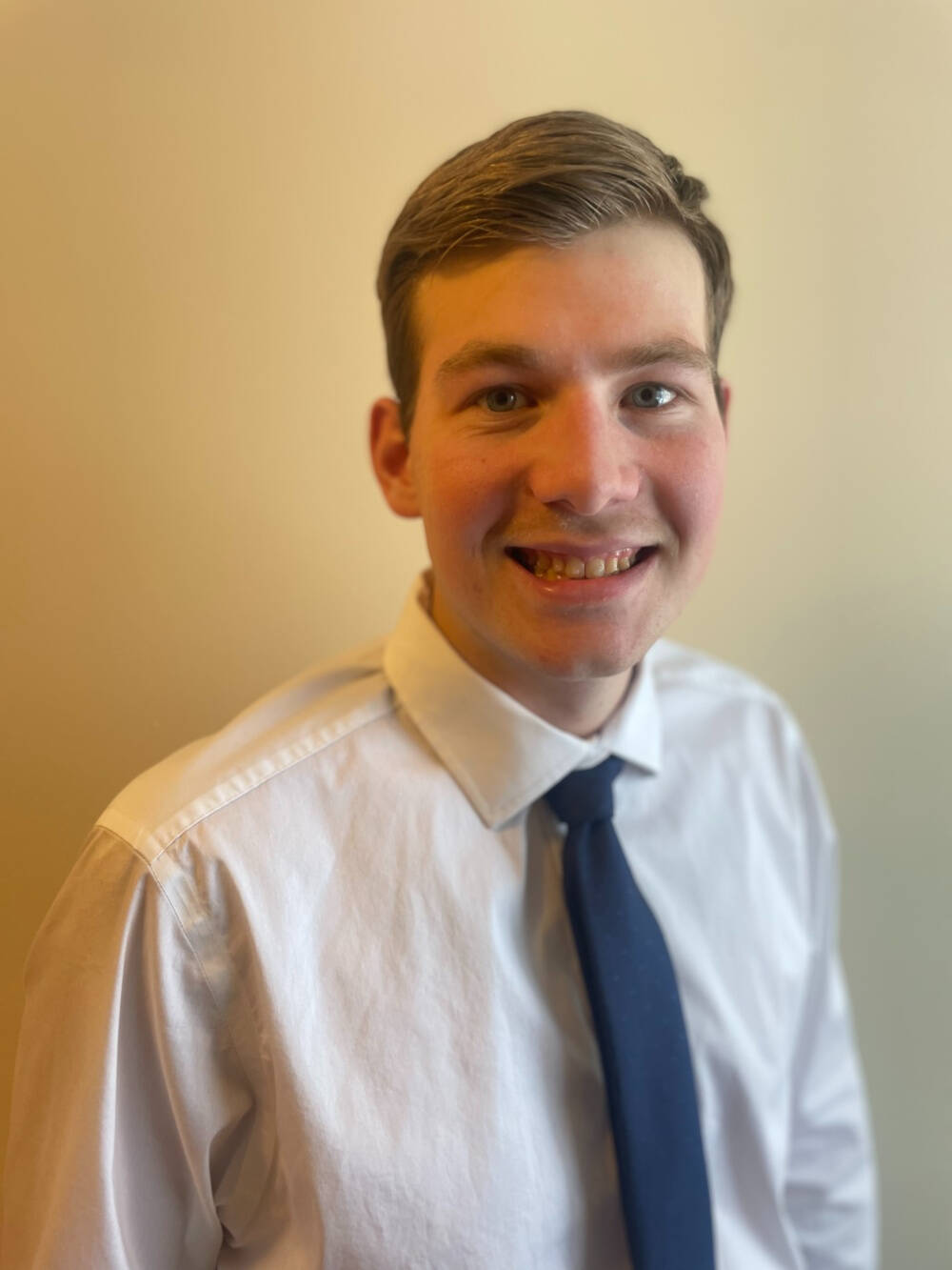 Colin Nagley in a white collared shirt with blue tie on a yellow backdrop.